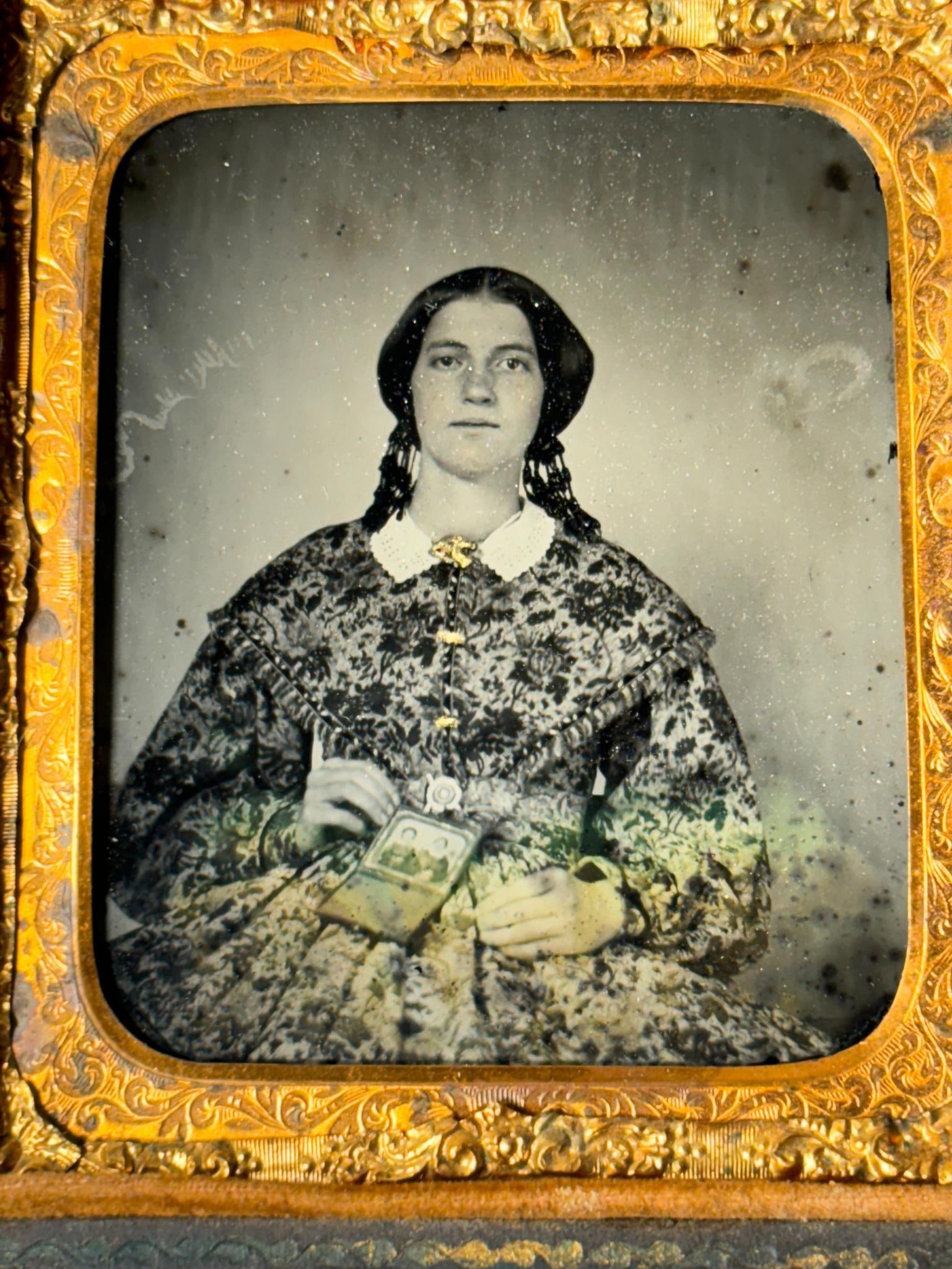 CIVIL WAR ERA AMBROTYPE - WOMAN HOLDING TINTYPE: Civil War period 1/6th plate ruby ambrotype of a seated woman wearing a beautiful floral print dress and holding a photograph on her lap. The cased hard image is open and shows a view of two women; mo