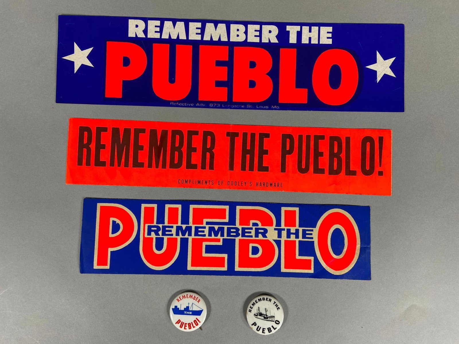 VIETNAM WAR ERA " REMEMBER THE PUEBLO " GROUPING: Vietnam era grouping of homefront "Remember The Pueblo" items including three bumper stickers and two buttons. One of the stickers is "Compliments of Dooley's Hardware" (Long Beach California) While t