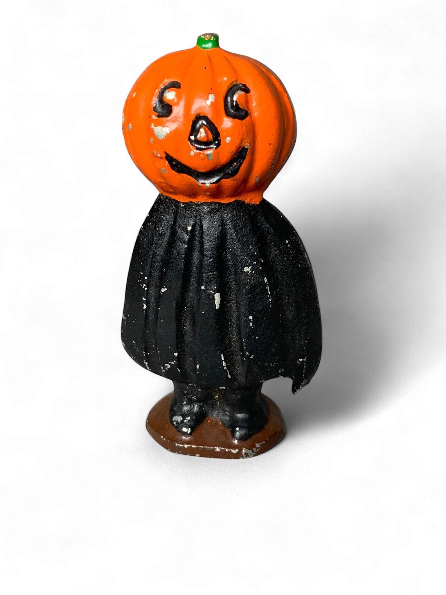 Vintage American Cast Aluminum Halloween Jack-o'-Lantern Figural: A vintage cast aluminum figural Halloween decoration, depicting a stylized individual with an orange jack-o'-lantern head and a black, possibly robed, body, standing on a brown octagonal base. The pie