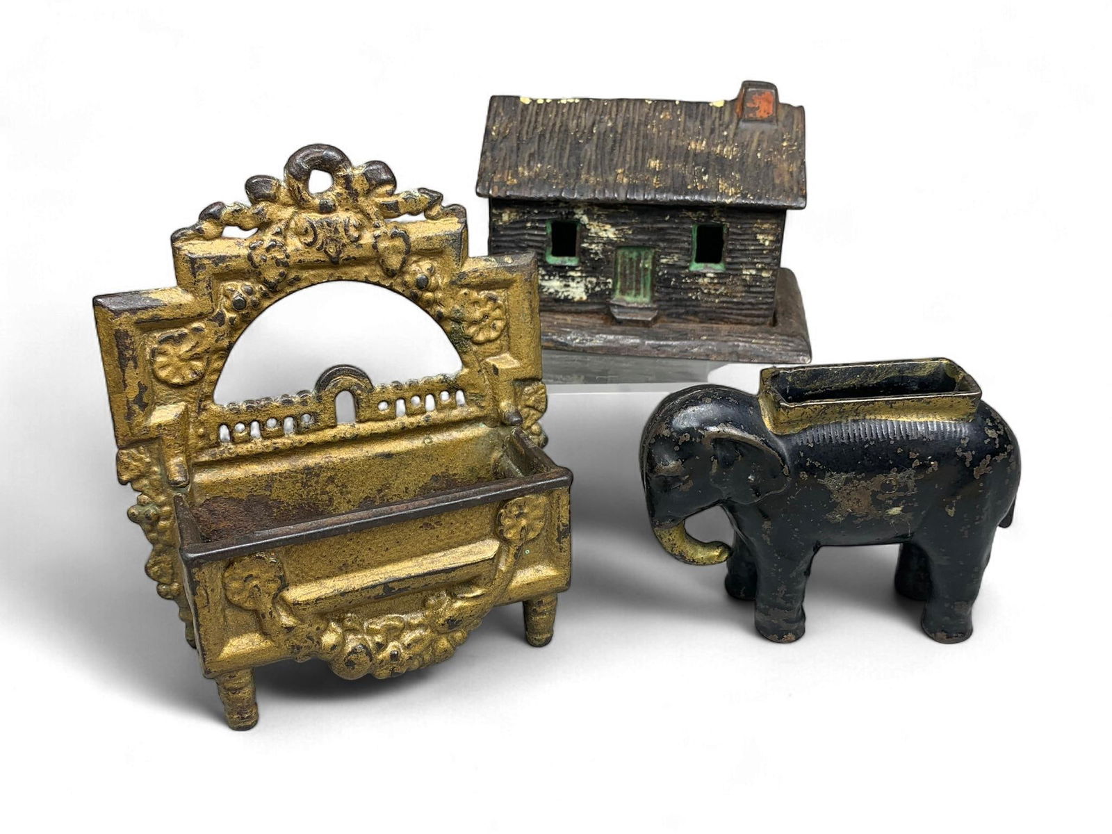 A Group of Antique Cast Iron Containers, including a Kyser & Rex Log Cabin Still Bank: A Kyser & Rex Log Cabin Still Bank, circa 1882, cast in iron with a rustic house-form. It features a hinged roof that opens to reveal a perforated interior. The bank retains remnants of dark, gold, gr