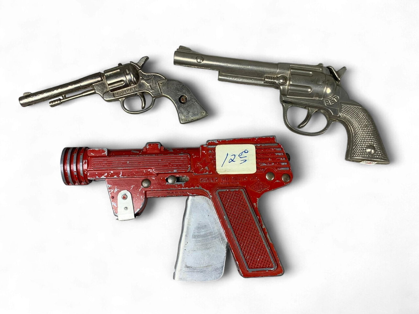 A Group of Vintage LMCO Super Nu-matic Jr. and Western-Style diecast Metal Toy Cap Guns: A lot comprising three vintage diecast metal toy cap guns. The primary piece is a red-painted, futuristic "ray gun" style model, identified through web intelligence as an LMCO Super Nu-matic Jr. Paper