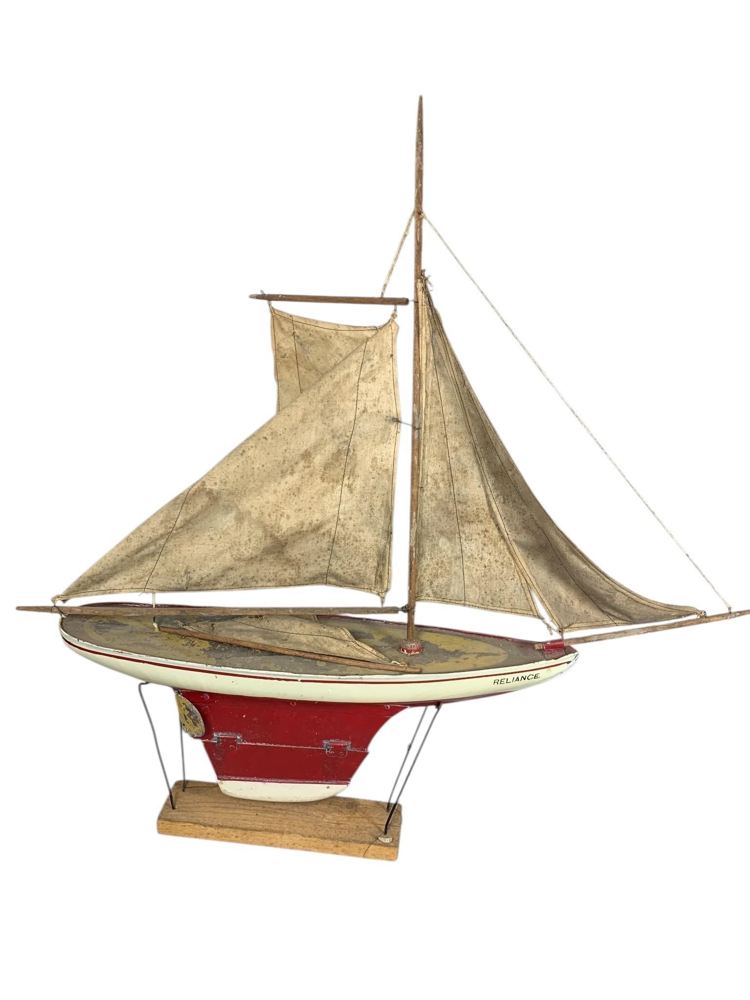 A Vintage Model Pond Yacht, 'Reliance': A vintage model pond yacht, named 'RELIANCE', styled as an early 20th-century racing yacht. It features a painted wooden hull, fabric sails, and wooden mast and spars. The hull is predominantly white