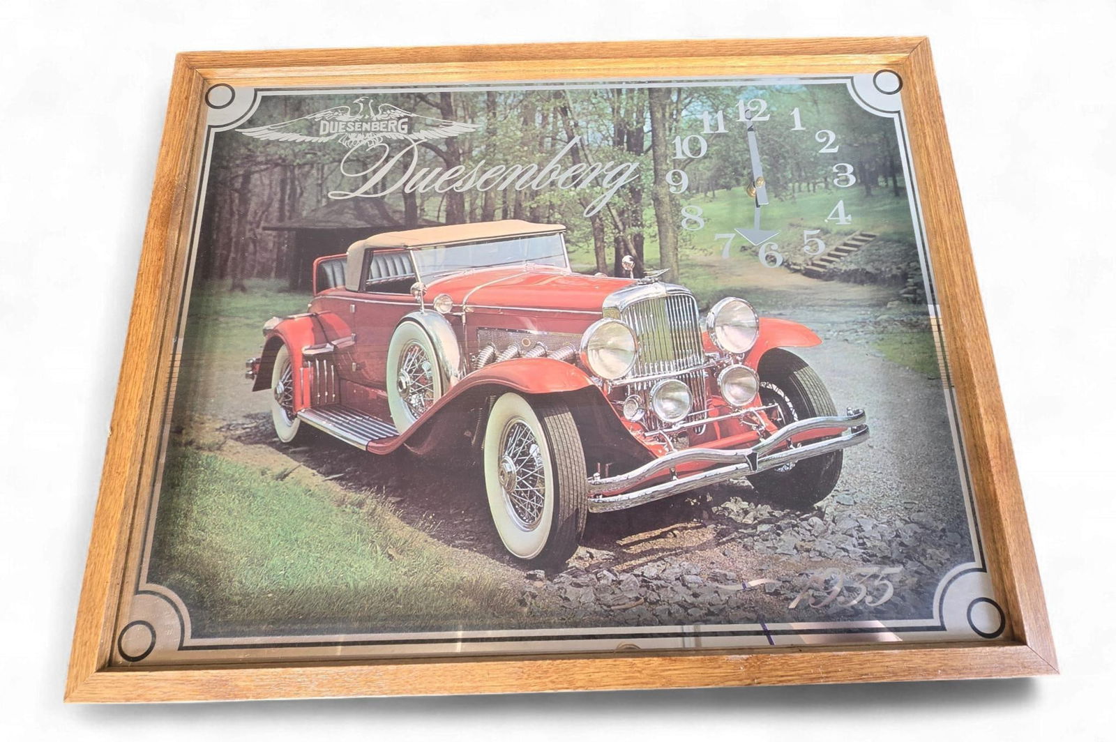 1935 Duesenberg Super Charged SJ Convertible Coupe Themed Wall Clock: A 1935 Duesenberg Super Charged SJ Convertible Coupe themed wall clock. This framed wall clock features a printed image of a classic red Duesenberg convertible against a wooded backdrop. The "DUESENBE