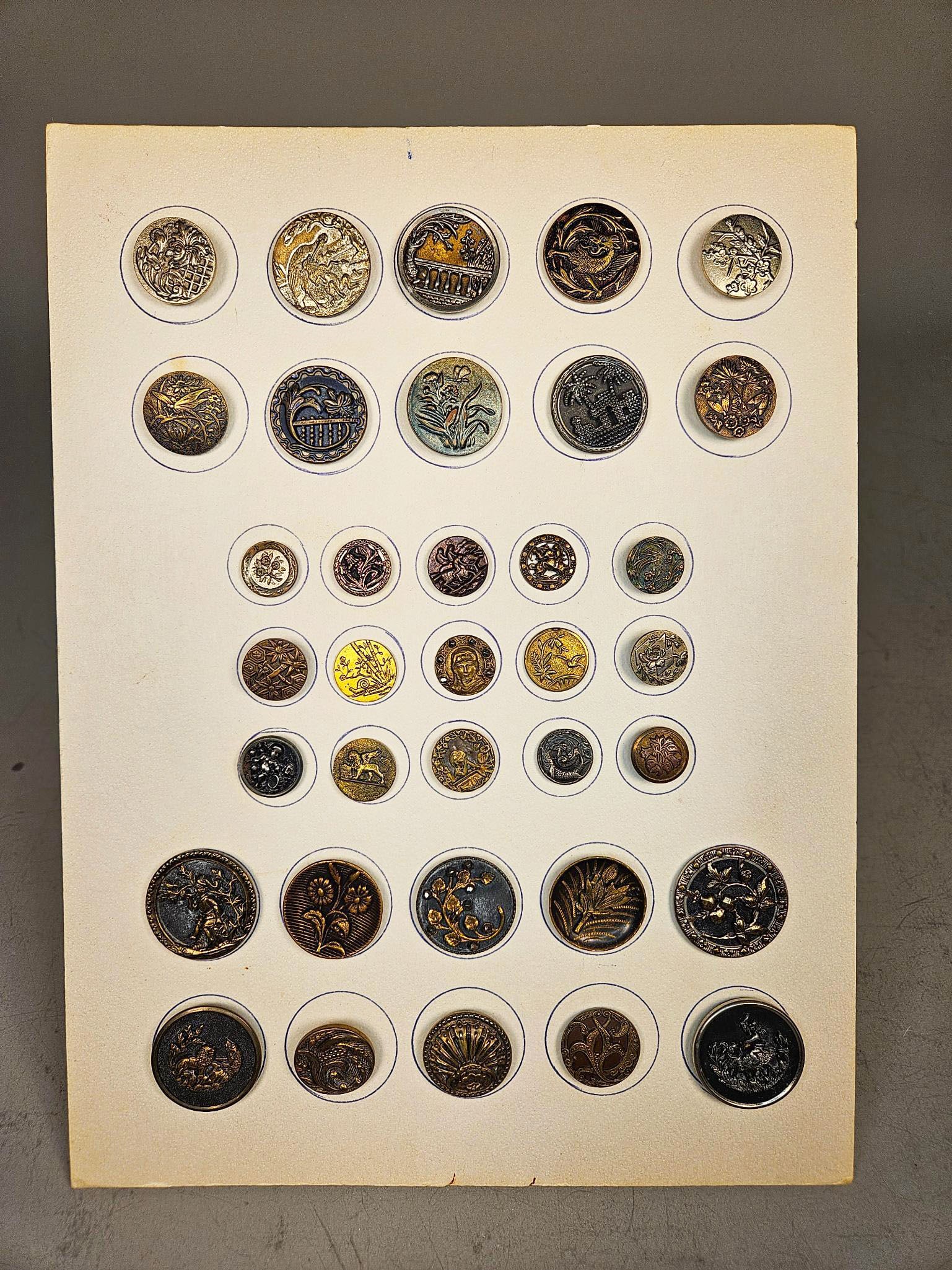 A Collection of Decorative Metal Buttons, Victorian to Art Nouveau: A collection of numerous decorative metal buttons, predominantly brass, bronze, and silver-toned alloys, meticulously arranged within blue-inked circles on a cream-colored mounting card. This diverse