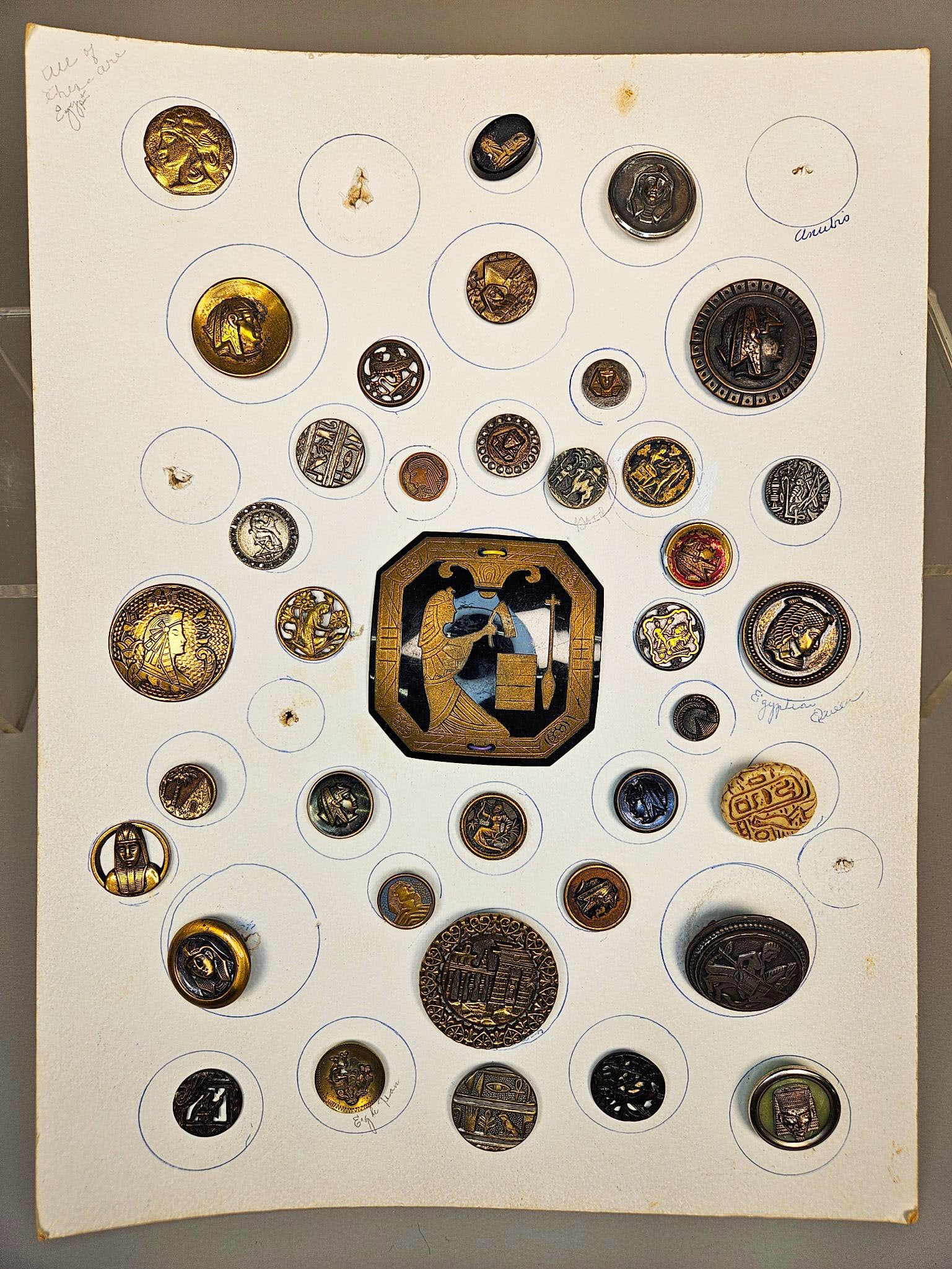 A Collection of Ancient Egyptian Revival and Other Decorative Buttons, May K Clark Collection: A curated group of decorative buttons, presented across group of white cardstock boards. The buttons predominantly feature an Ancient Egyptian Revival style, depicting pharaohs, queens, sphinxes, deit
