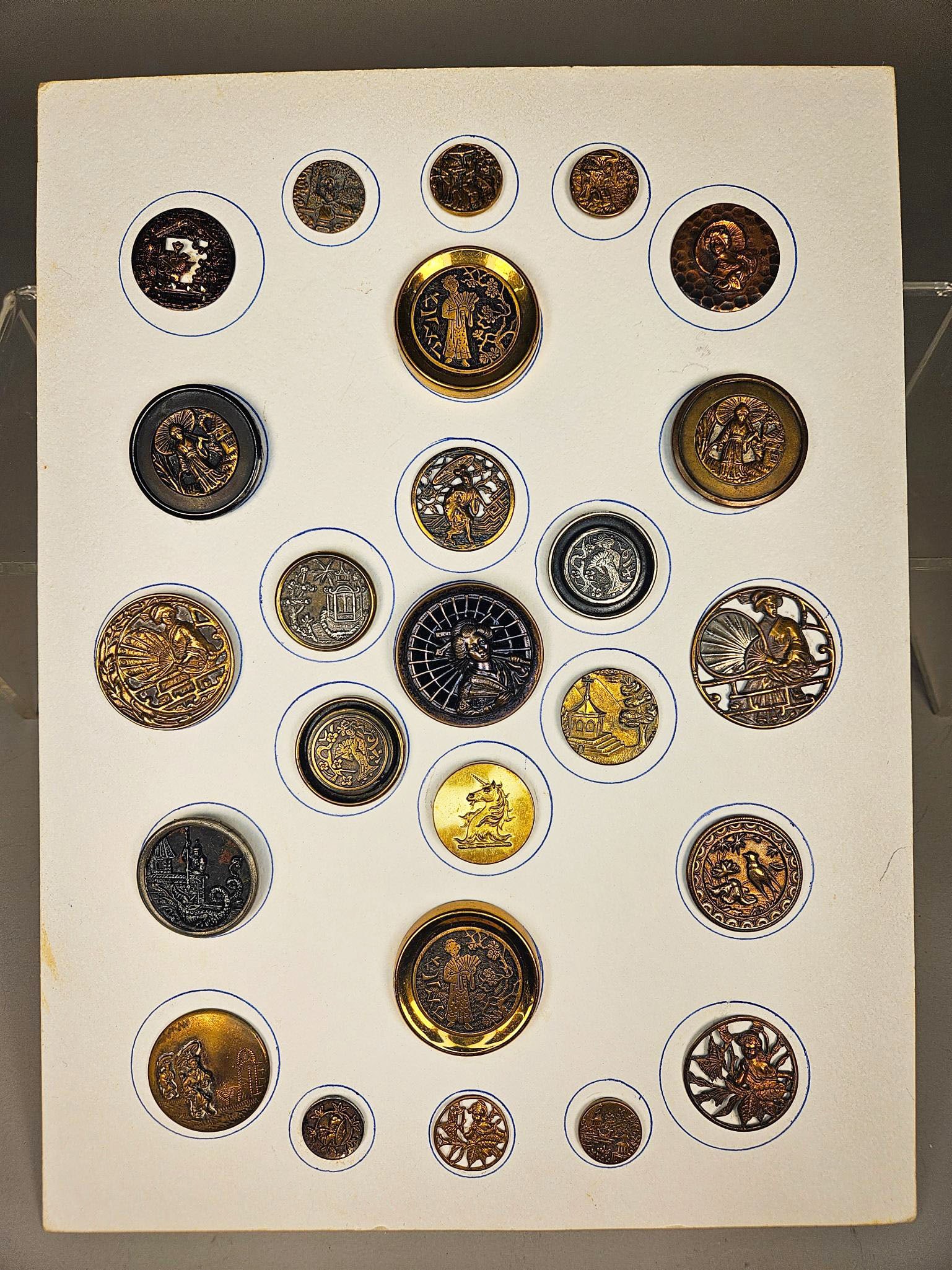 A Collection of Victorian Japonesque Metal Picture Buttons: A group of decorative metal buttons, predominantly circular and varying in size, meticulously mounted on a white display board with blue-inked outlines. The buttons exhibit a diverse range of styles r