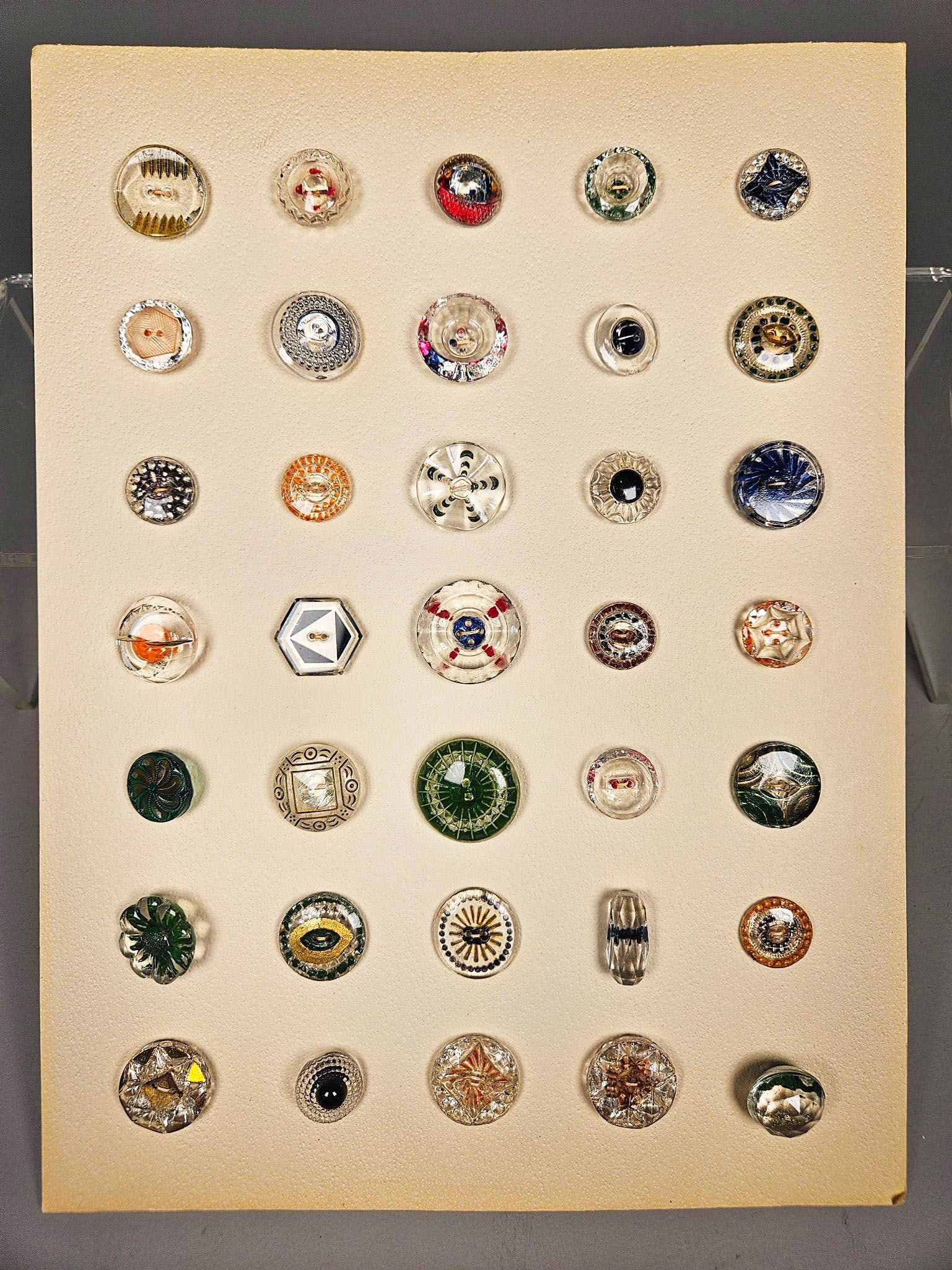 A Collection of Assorted Decorative Glass Buttons: A group of decorative buttons, primarily crafted from clear and colored glass, presented on an off-white display card. The buttons showcase a diverse array of styles, including molded facets, internal