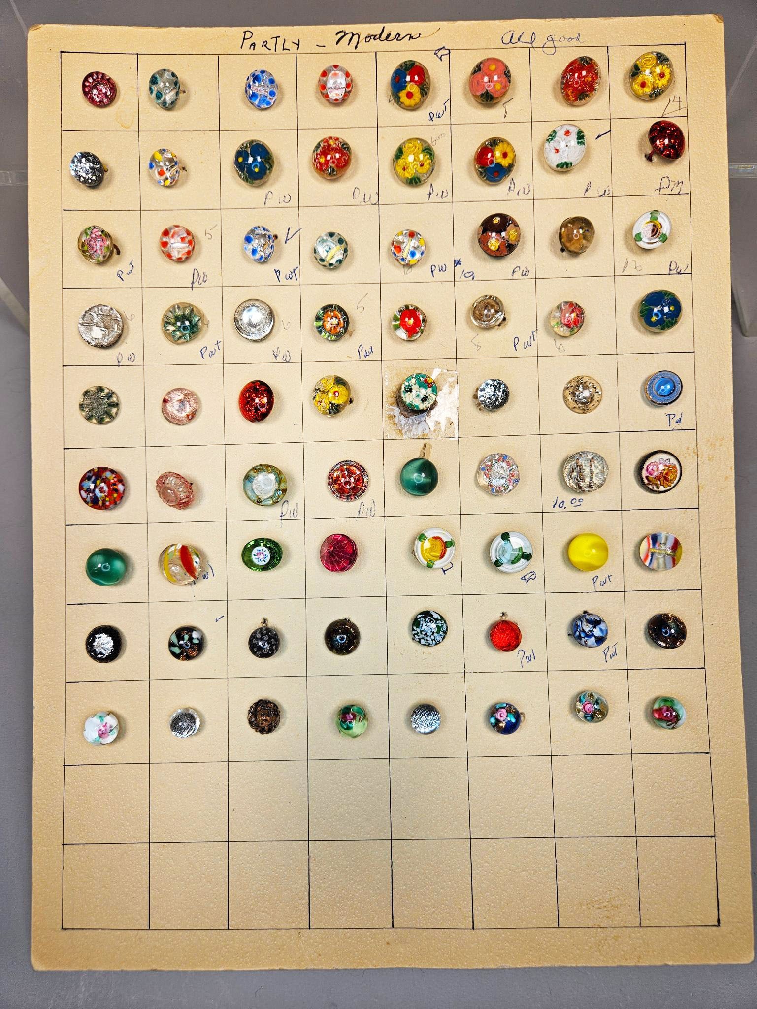 A Collection of Vintage and Modern Art Glass Buttons: A collection comprising group of individual glass buttons, predominantly spherical or domed, meticulously arranged and secured within a grid pattern on an aged, cream-colored card display. The buttons