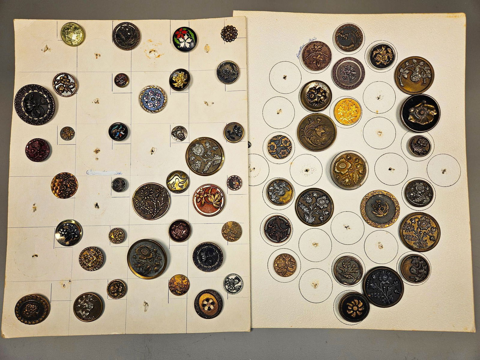 A Collection of Late 19th to Early 20th Century Metal Buttons, Featuring Botanical, Floral, and: A group of antique or vintage buttons, predominantly circular and varying in size, displayed across group of off-white cardboard sheets. The buttons exhibit a diverse range of styles, heavily featurin