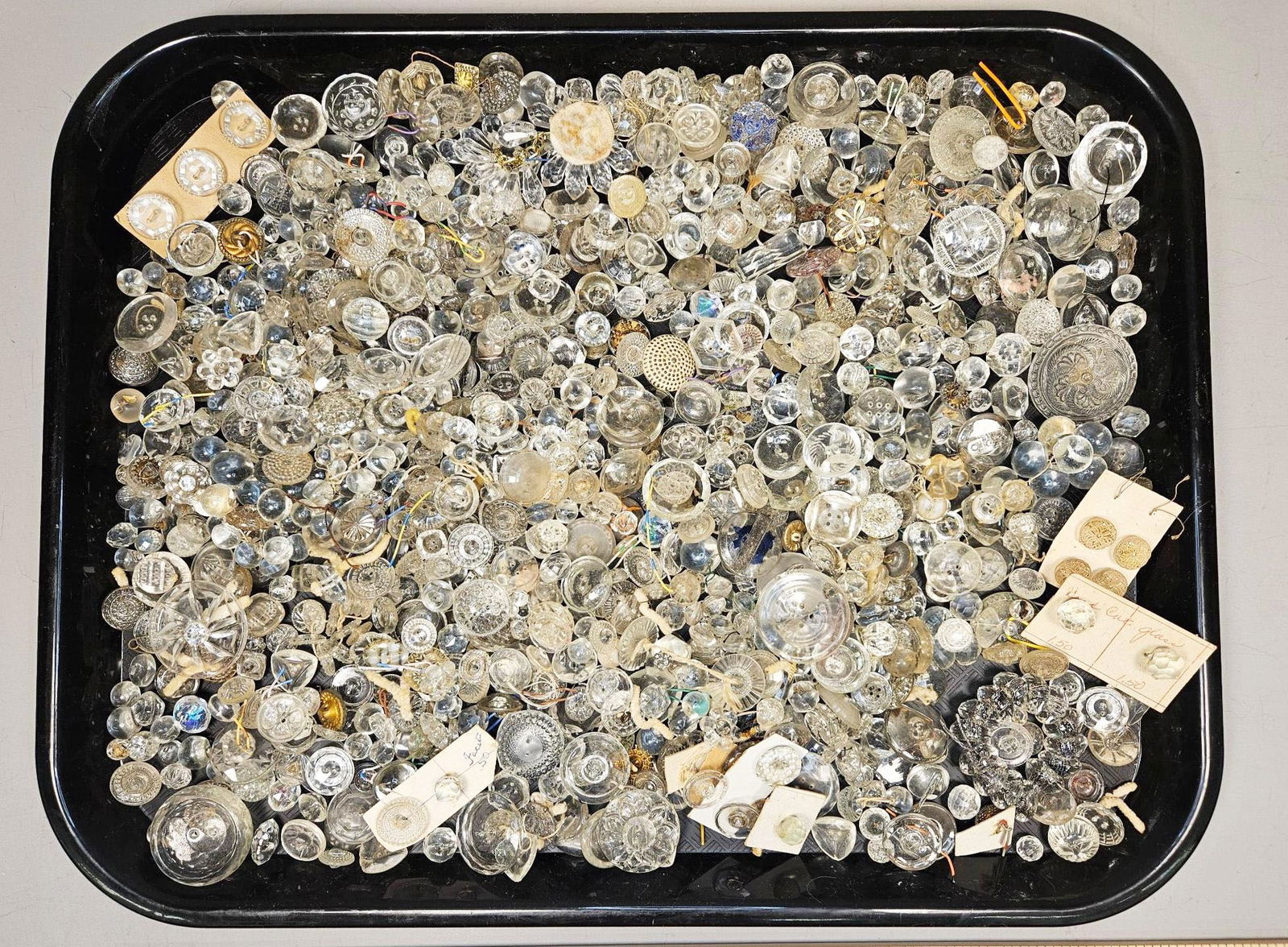 A Large Collection of Varied Clear Glass Buttons, Some Hand-Cut: A large collection of buttons, primarily composed of clear and colorless glass, presented in a black tray. The collection displays a significant variety in style, period, and form, encompassing facete