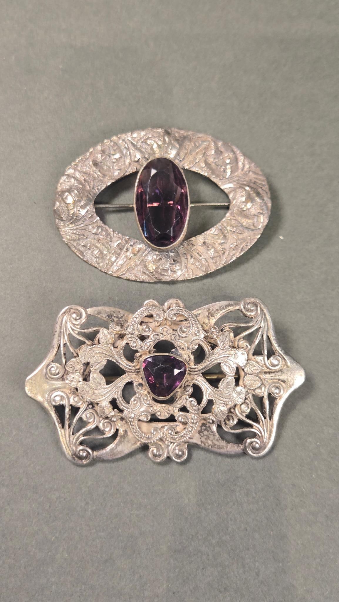 A Pair of Victorian Brooches: A pair of Victorian-style brooches. Both pieces are constructed from a silver-toned metal. Each brooch features a central, faceted purple stone; one is an oval cut, while the other presents as a shiel