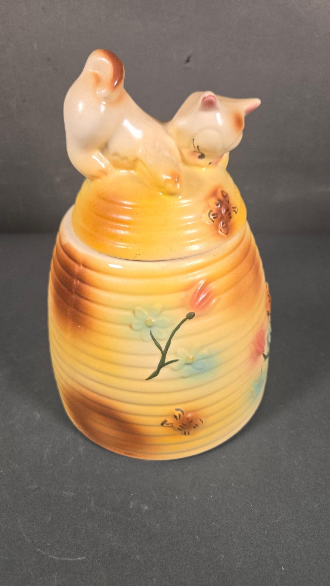 American Bisque Cat Kitten On Beehive Flower Cookie Jar, circa 1950s: An American Bisque 'Cat Kitten On Beehive Flower' cookie jar, dating to the circa 1950s. This lidded glazed ceramic jar features a horizontally ribbed, predominantly yellow body with brown gradient ac