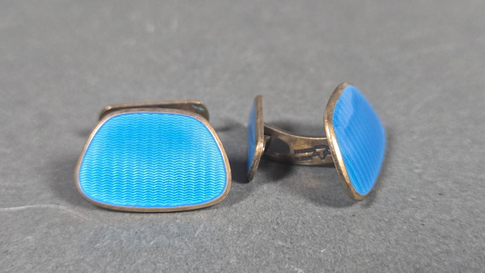 A Pair of Sterling Silver and Blue Enamel Cufflinks, Made in Norway: A pair of sterling silver cufflinks featuring bright blue enamel work. The primary face of each cufflink is a rounded, trapezoidal shape with a wavy guilloche pattern under translucent blue enamel. Th