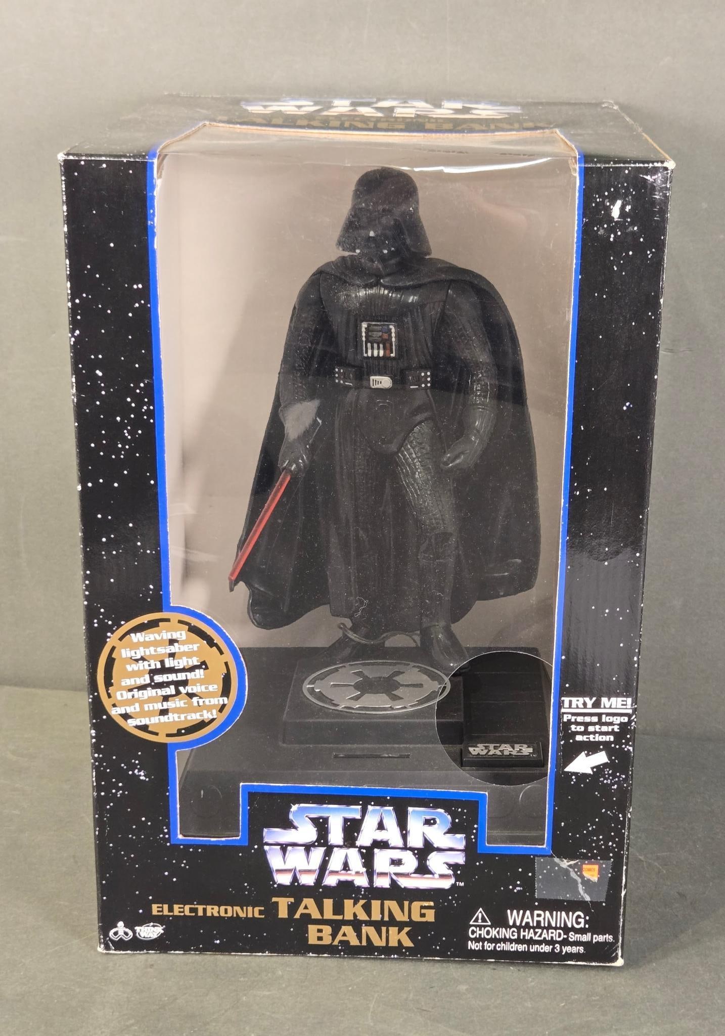Star Wars Darth Vader Electronic Talking Bank by Thinkway Toys, 1996: A Star Wars Darth Vader Electronic Talking Bank, manufactured by Thinkway Toys in 1996. The item features a plastic Darth Vader figure, clad in black and holding a red lightsaber, standing on a base w