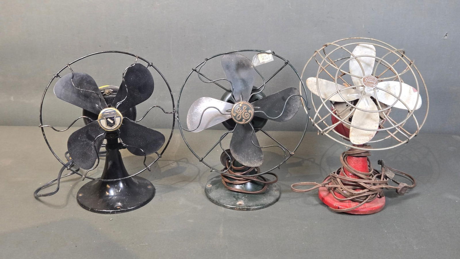 Three Vintage Metal Electric Desk Fans, including General Electric, Graybar, and Young examples: A lot comprising three vintage metal electric desk fans, likely from the early to mid-20th century, each featuring a distinct design and finish. The leftmost fan is a black-painted metal model with fo