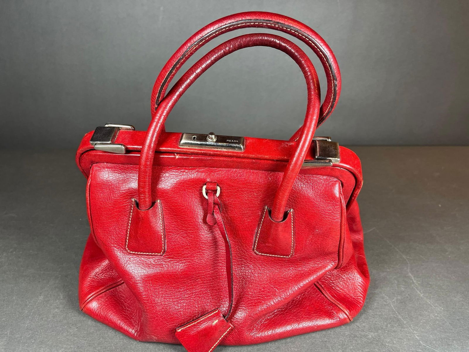 A Prada Red Pebbled Leather Doctor's Bag Handbag: A Prada red pebbled leather handbag, designed in a doctor's bag style with a structured frame opening and dual rolled top handles. It features silver-tone hardware, notably a lock mechanism engraved w