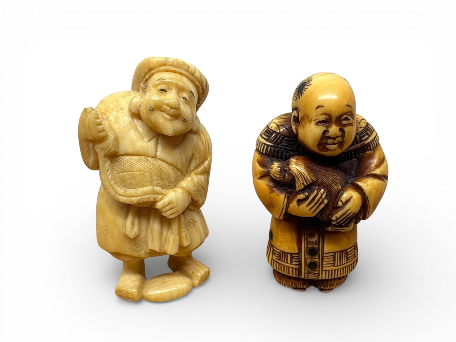 Carved Bone Japanese Netsuke Figures: Japanese carved netsuke figures, crafted from a bone-like material with varying patination. The lighter-toned figure depicts a jovial, robed elderly man wearing a cap. The darker, more richly stained