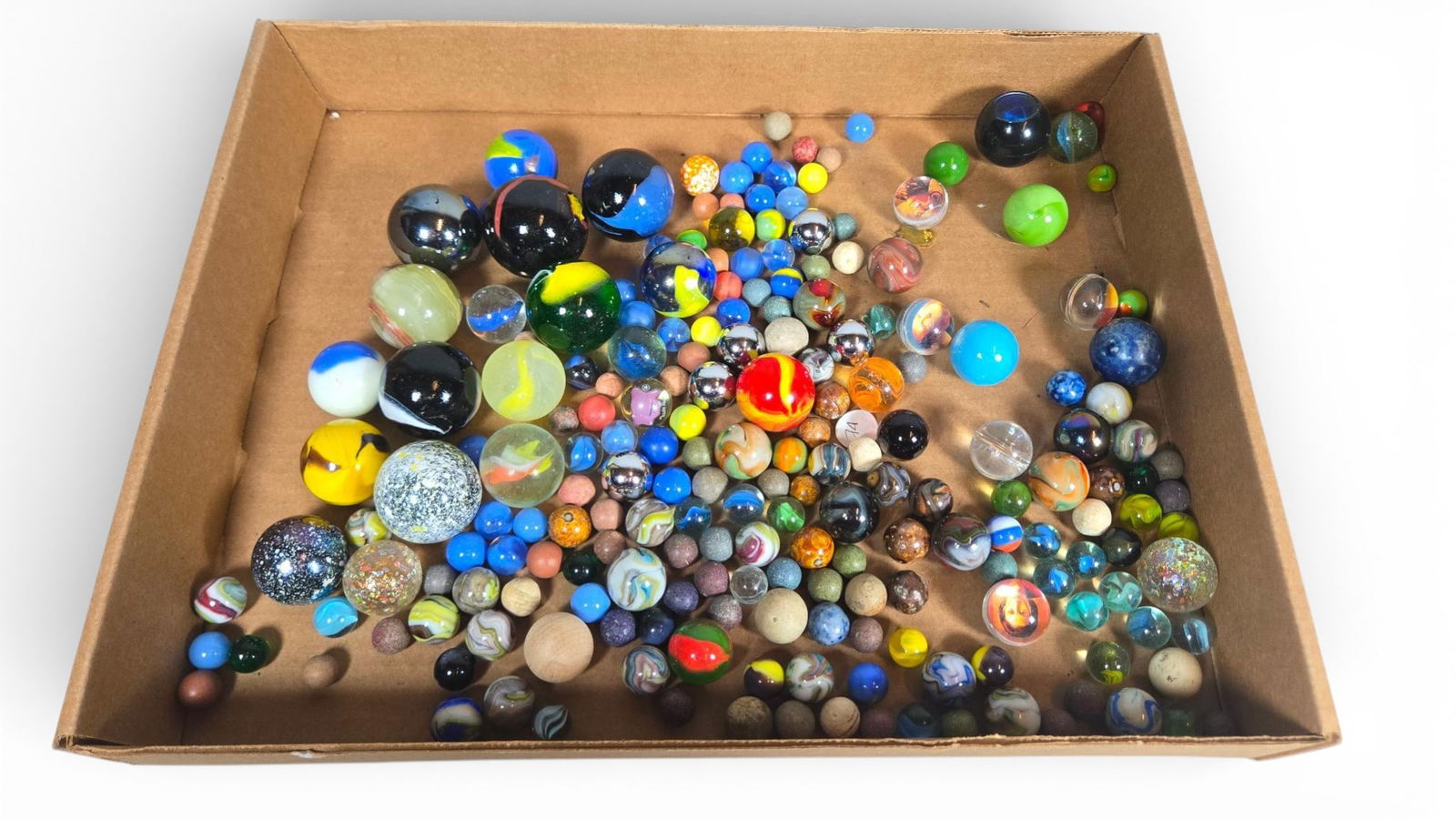 Collection of Assorted Vintage and Contemporary Marbles: Collection of or more assorted marbles spanning mixed eras and materials, densely packed within a corrugated cardboard box. The collection includes spheres constructed from glass, ceramic, stone, and