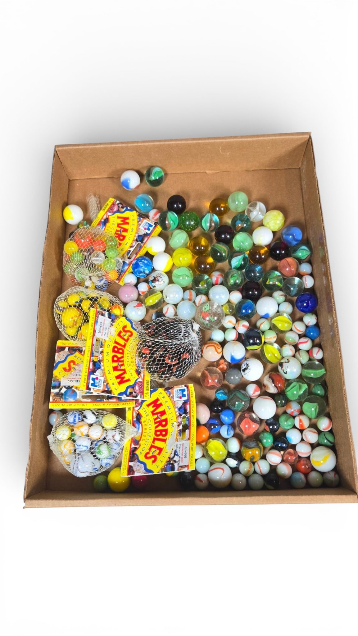 Assortment of Machine-Made Glass Marbles with Premium Collector Series Sets: The collection comprises a large assortment of machine-made glass marbles contained within a brown cardboard storage box. The lot includes individually packaged groups of marbles, each sealed in a