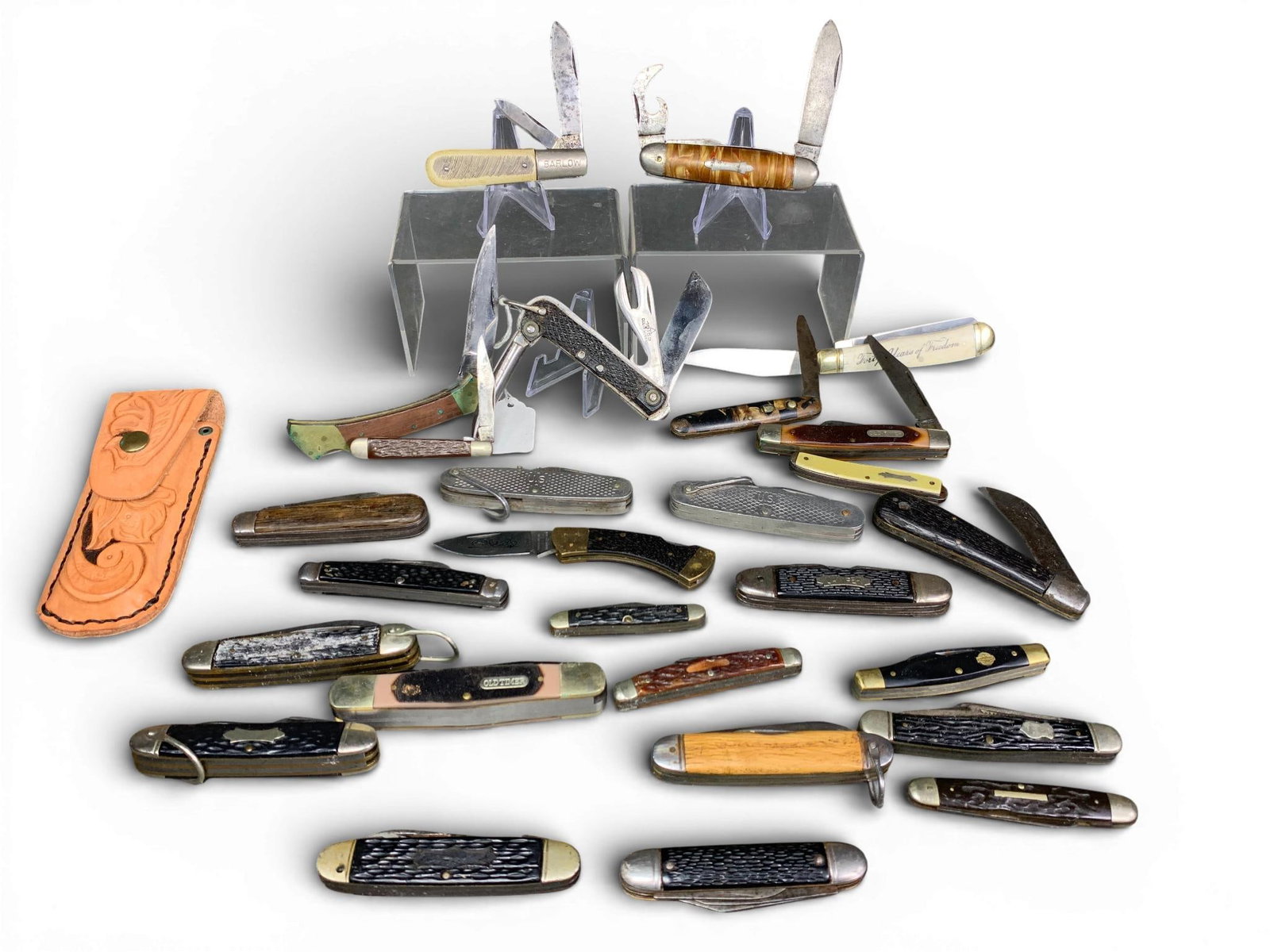 A Collection of Vintage and Modern Pocket Knives with Tooled Leather Sheath: A collection of various vintage and modern folding pocket knives presented with a single tooled tan leather belt sheath. The assortment includes multiple styles such as multi-tool camper knives, Barlo