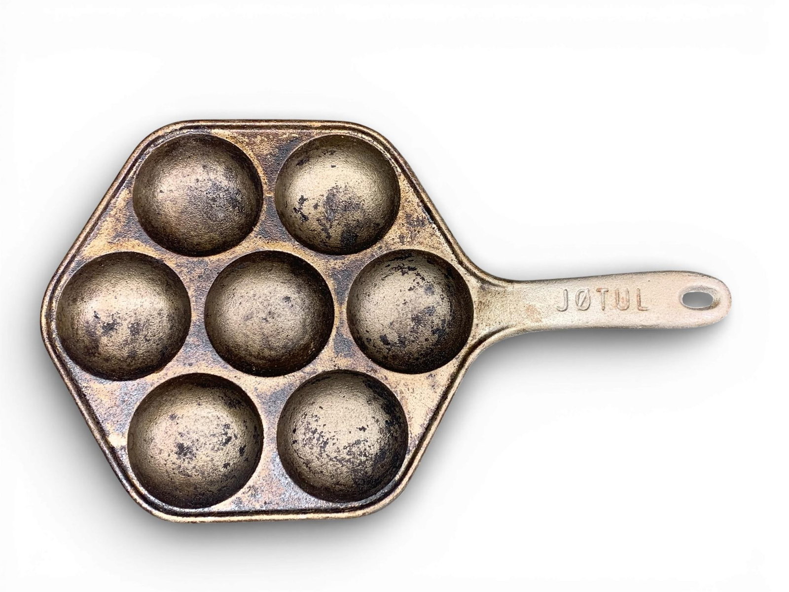 Jøtul Cast Iron Aebleskiver Pan: A Jøtul cast iron aebleskiver pan featuring a hexagonal frame with seven spherical cooking wells. The piece is constructed of cast iron and includes a handle with a hanging hole. The topside of the h