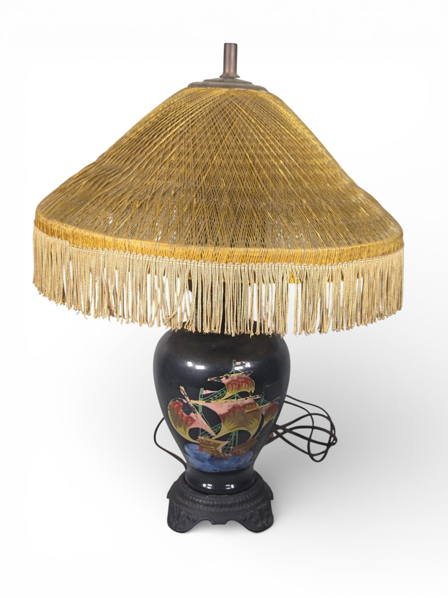 Mid-20th Century Ceramic Table Lamp with Nautical Motif and Fringed Cord Shade: This mid-20th-century table lamp features a tapered ceramic body with a black glaze, decorated with a stylized, multi-colored sailing vessel motif. The lamp is mounted on a heavy, scalloped cast-metal