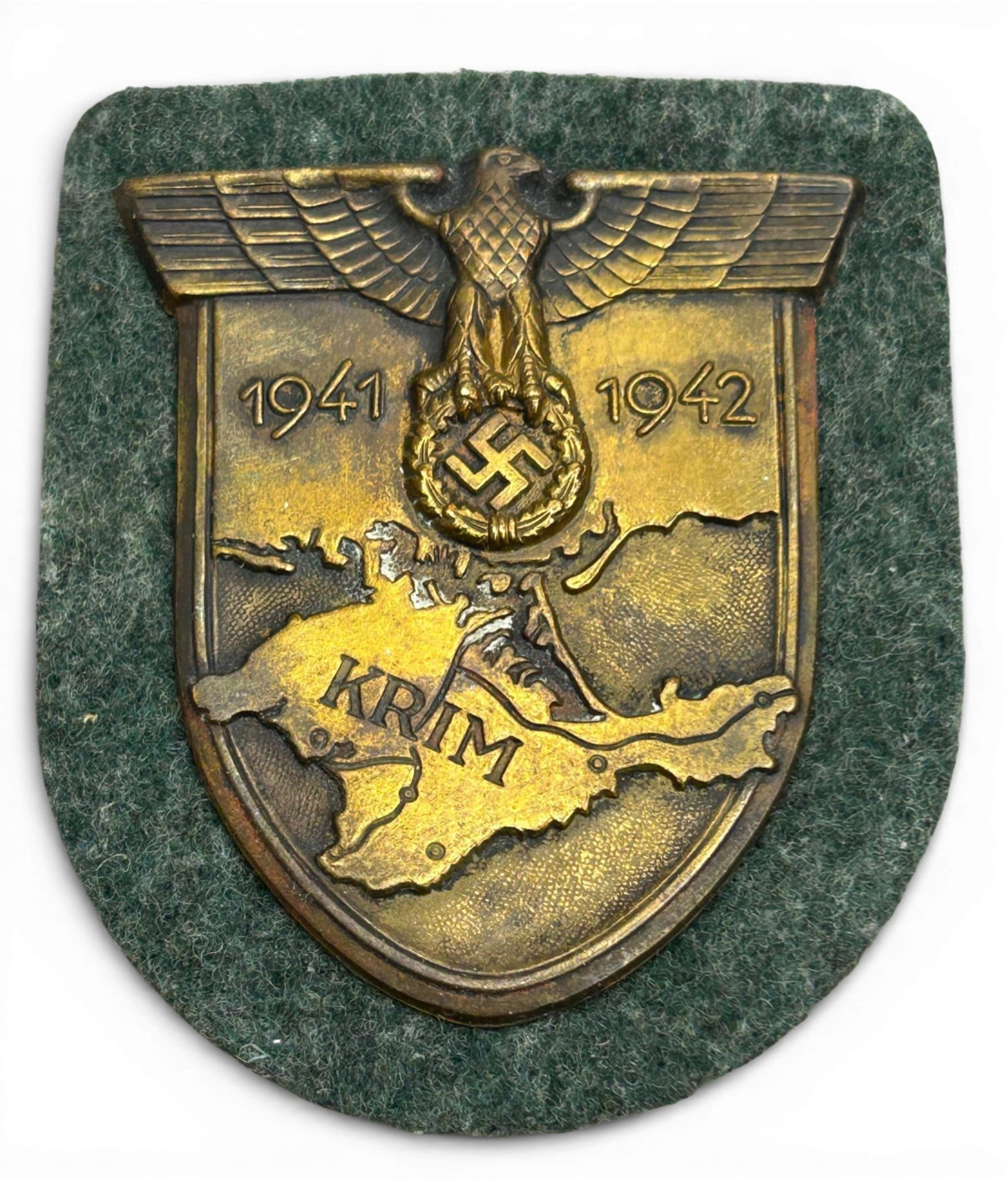 WWII Nazi German Krim Shield: German WWII Army Heer 1941 – 1942 KRIM Sleeve Shield. Measures 2.5" x 2.875”. Mounted onto an OD green wool backing. Has a steel back plate and 4 attachment prongs under the tan paper on the r