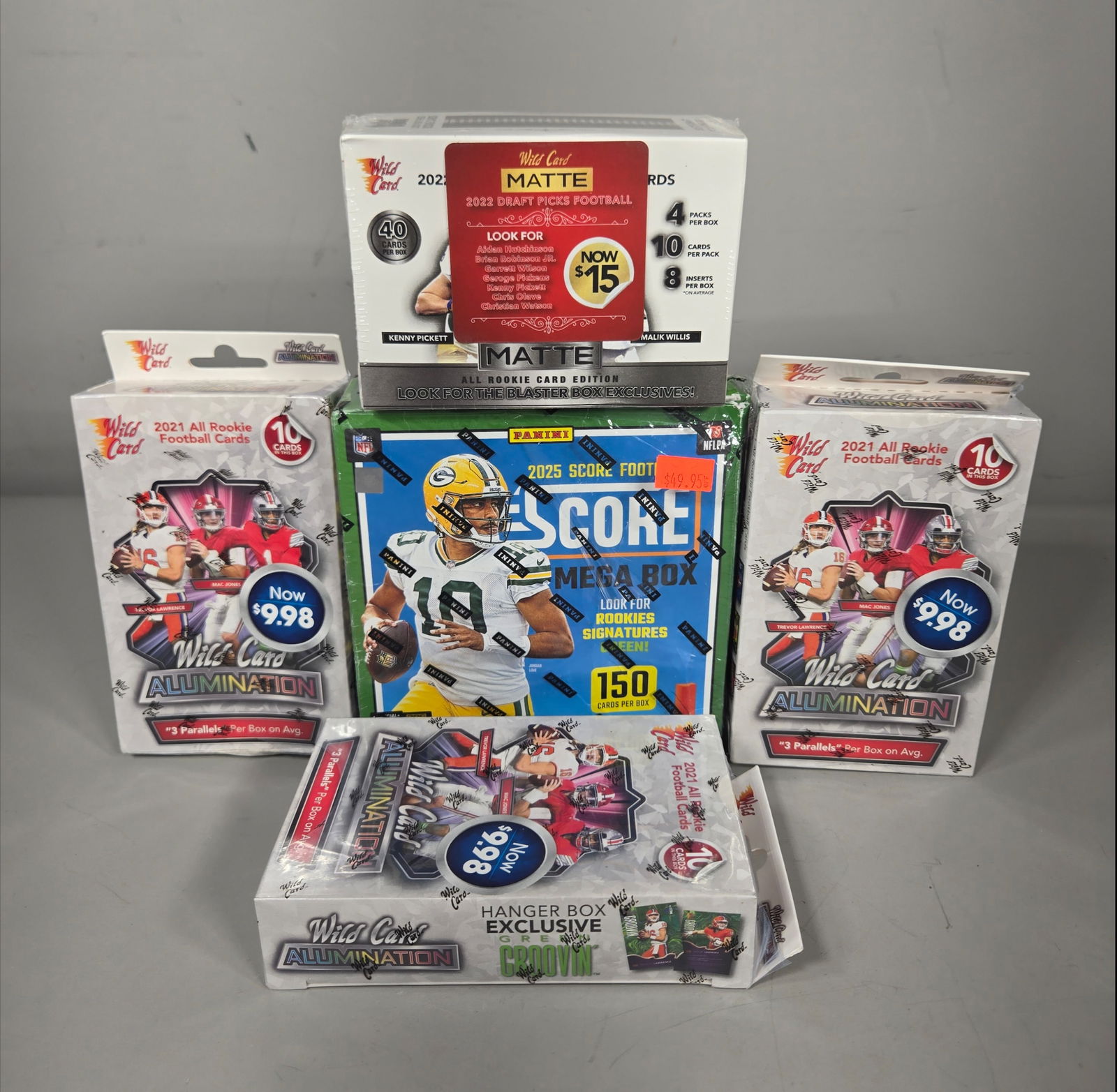 A Collection of Panini Score and Wild Card Alumination Football Trading Card Boxes: A collection of factory-sealed modern football trading card boxes. The assortment consists of one 2025 Panini Score Mega Box, one 2022 Wild Card Matte Draft Picks Football blaster box, and three 2021