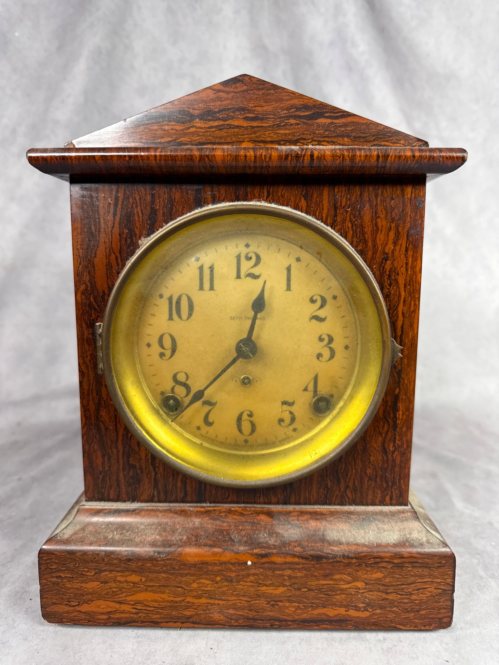 Seth Thomas Red Adamantine Mantle Clock: A Seth Thomas mantle clock featuring a rectangular wooden case finished in a reddish-brown marbled Adamantine veneer. The circular dial is protected by a glass panel within a metal bezel and displays