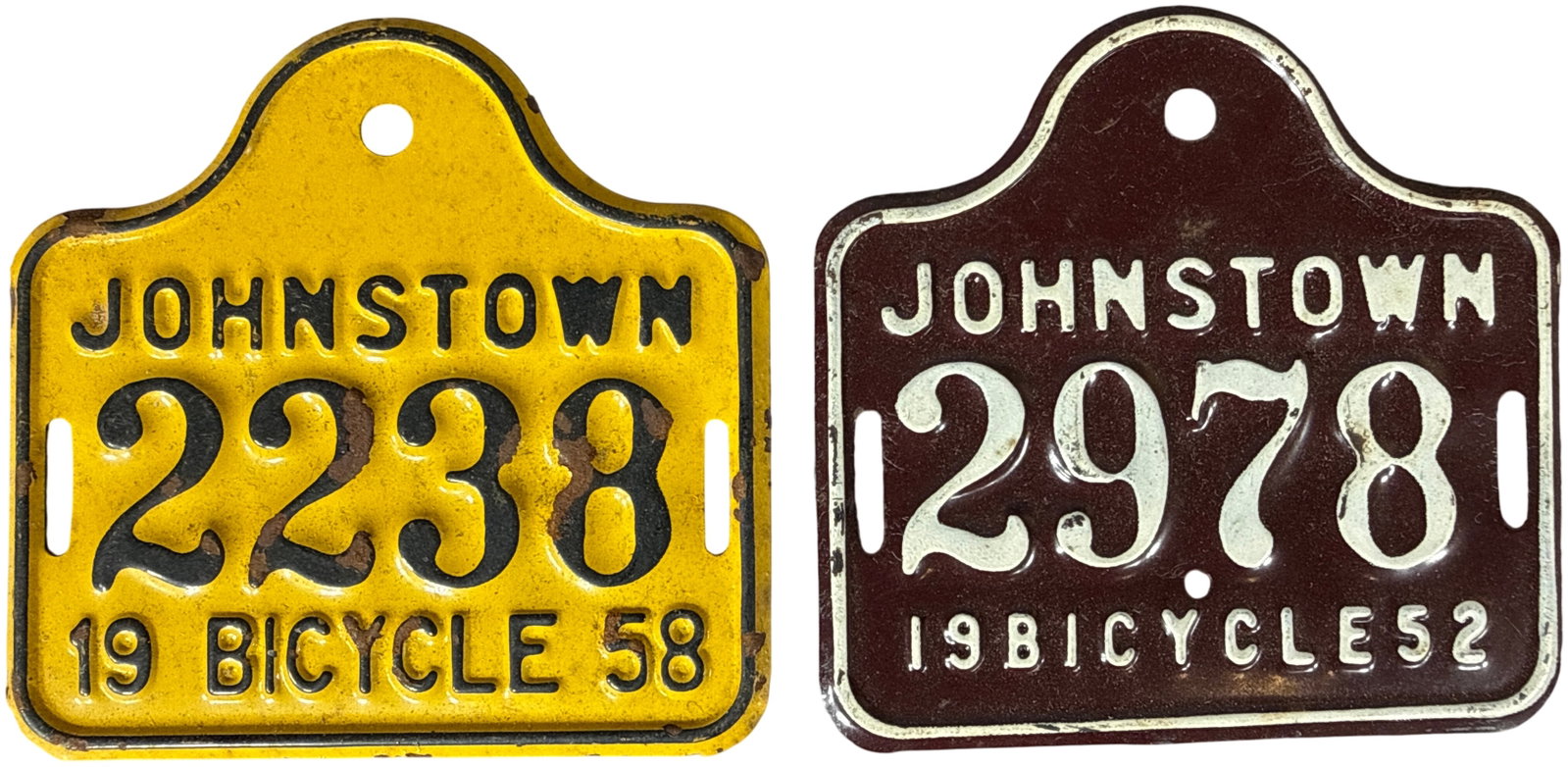 Vintage Johnstown Embossed Metal Bicycle License Plates, 1952 and 1958: This lot features municipal bicycle registration tags from Johnstown. The plates are constructed of embossed painted steel. The 1952 registration tag features a maroon background with cream-colored em