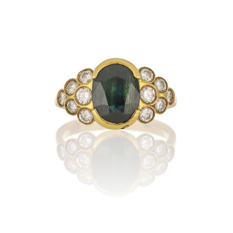 Anello in oro giallo con zaffiro australiano: Anello in oro giallo con zaffiro australiano taglio ovale, ct. 5,00 ca. e 12 diamanti taglio brillante disposti a formare due triangoli ai fianchi, per ct. 0,50 ca. Misura n. 13. Peso 6,66 g.
