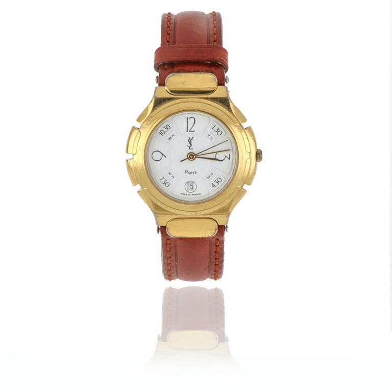 YVES SAINT LAURENT Orologio da donna al quarzo in: YVES SAINT LAURENT Orologio da donna al quarzo in metallo dorato, con quadrante bianco, piccolo datario ad ore 6. Non testato. Scatola e fibbia originale.
