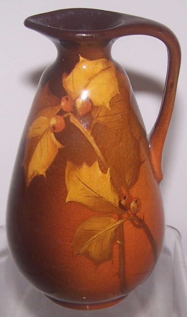 *ROOKWOOD ART POTTERY CRUET: Having standard glaze and decorated with holly leaves and berries on a ground shaded brown, green and rust, marks include impressed Rookwood flame logo indicating date 1899, shape #746 D, signed indis