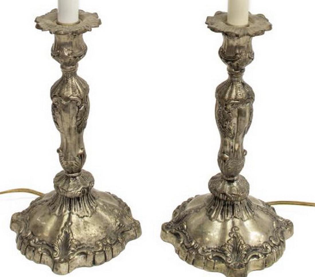 (2) DECORATIVE SILVER-TONE METAL CANDLESTICK LAMPS: (pair) Decorative silver-tone metal table lamps, 20thc., each fashioned as a rocaille and foliate decorated candlestick, with cylindrical stem leading to a single socket, one lacking finial at harp, a