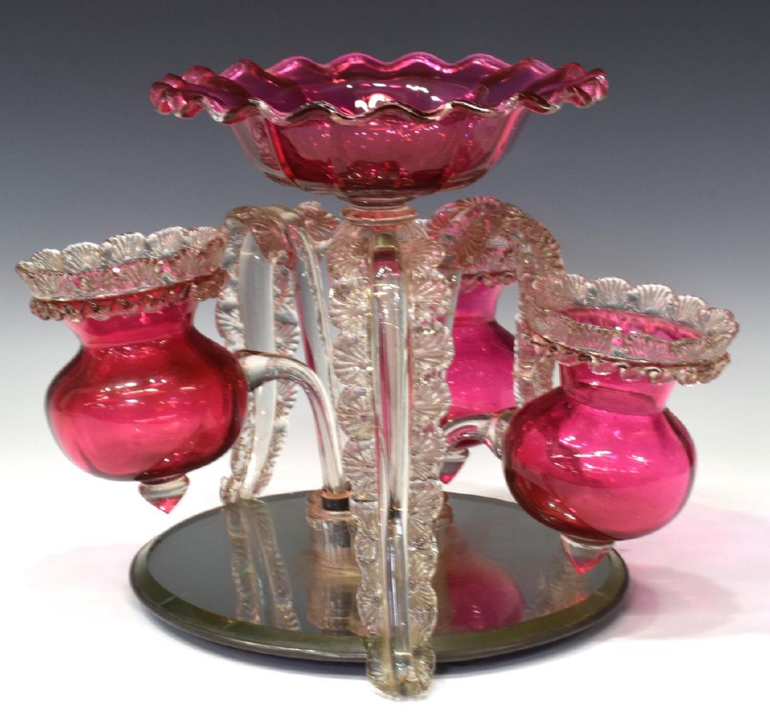 CRANBERRY & COLORLESS ART GLASS CENTERPIECE: Cranberry and colorless glass centerpiece on mirror, assembled, the ruffled bowl centering three smaller bowls with applied shell rim and three elongated foliate form accents, rising on a circular mir