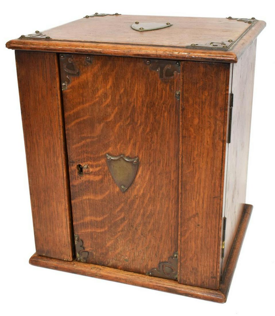 ENGLISH EDWARDIAN METAL-BOUND OAK TOBACCO CABINET: English Edwardian oak tobacco cabinet, early 20th c., fancy metal bindings and shield-form plaques on the case, opening to fitted interior with three drawers, spring mechanism opening side panel, able