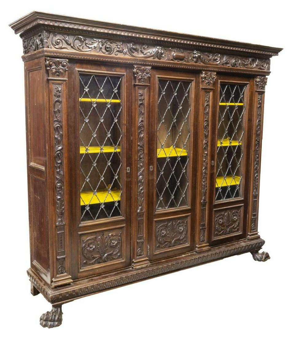 ITALIAN RENAISSANCE REVIVAL WALNUT BOOKCASE: Italian Renaissance Revival walnut bookcase, early 20th c., molded cornice, over grotesque and foliate carved frieze, three glazed doors with iron lattice overlay, ionic pilasters, rising on paw feet,