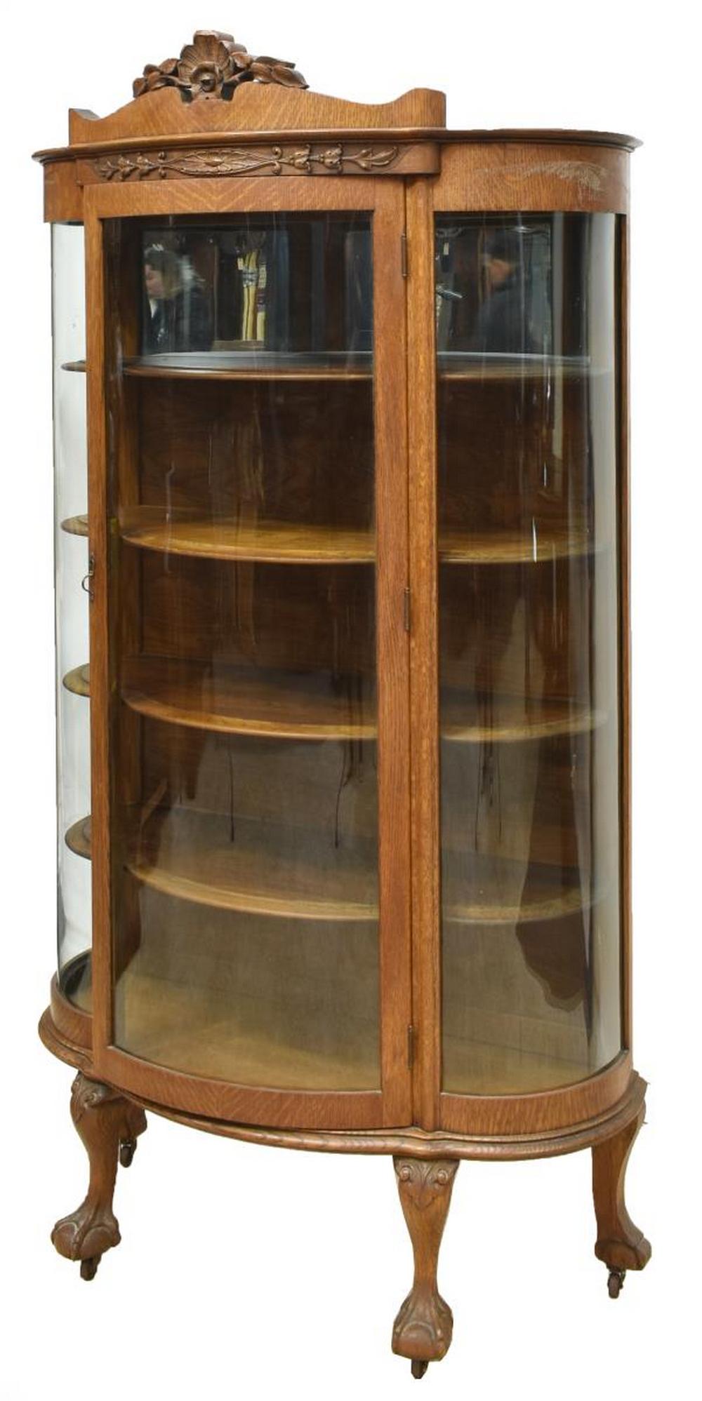 AMERICAN OAK CURVED GLASS CHINA CABINET, 20TH C: American oak curved glass china cabinet, 20th c., a central cornice with applied foliate carving, curved glass door and side panels, shelved interior with an upper mirrored panel, carved paw feet risi