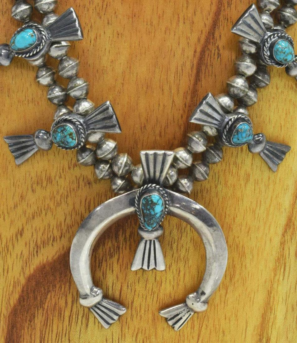NATIVE AMERICAN TURQUOISE SQUASH BLOSSOM NECKLACE: Native American silver (content unknown) squash blossom necklace, bench bead strand, twelve blossoms, central naja, each with a bezel-set turquoise cabochon, approx 25.25"l, naja: approx 2 5/8"h, 2 1/