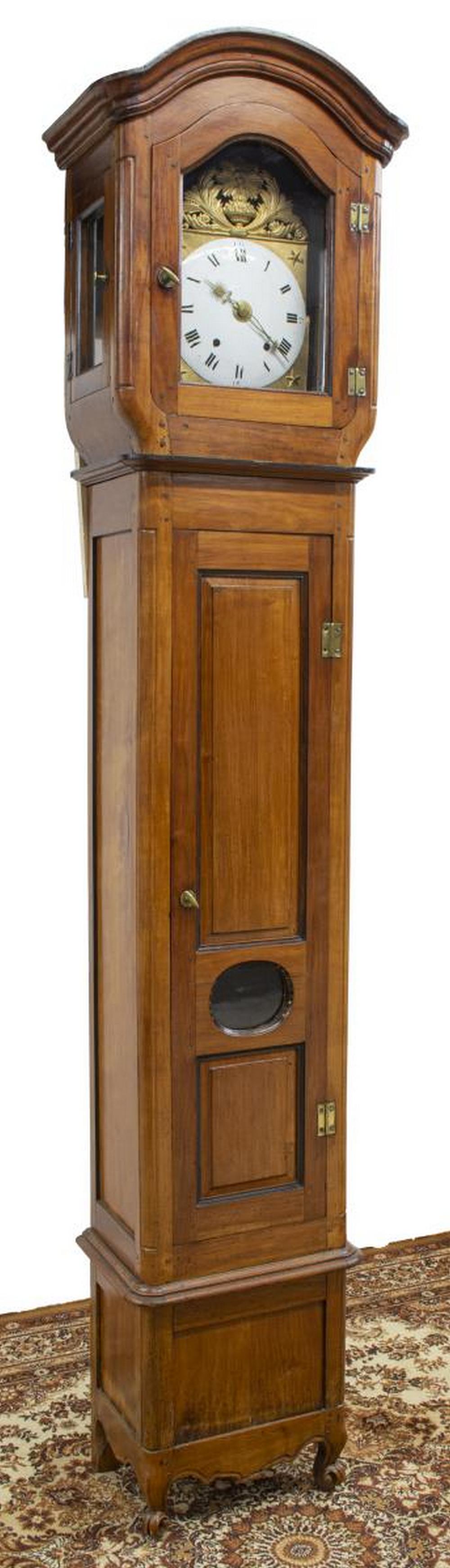 FRENCH PROVINCIAL FRUITWOOD LONGCASE CLOCK: French Provincial Louis XV style fruitwood longcase clock, 19th c., arched molded cornice, glazed inspection doors and pendulum view, housing gilt metal repousse frame depicting starburst backed mask 