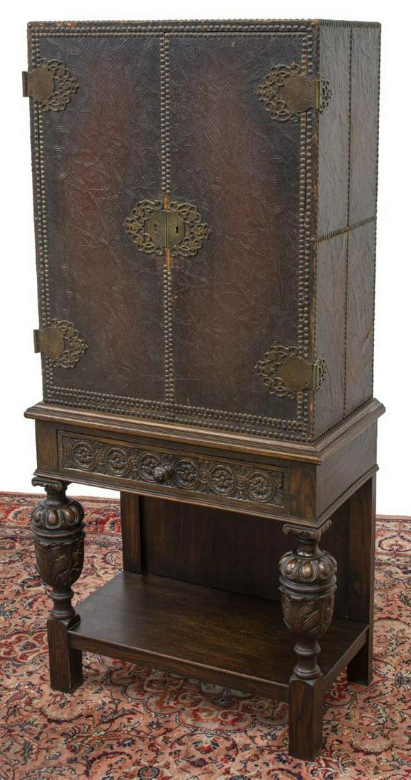 SPANISH RENAISSANCE REVIVAL LEATHER CABINET STAND: Spanish Renaissance Revival oak cabinet on stand, late 19th c., leather-clad upper case with double doors, elaborate scrolled hinges and escutcheon, interior shelves of varied size, over single frieze