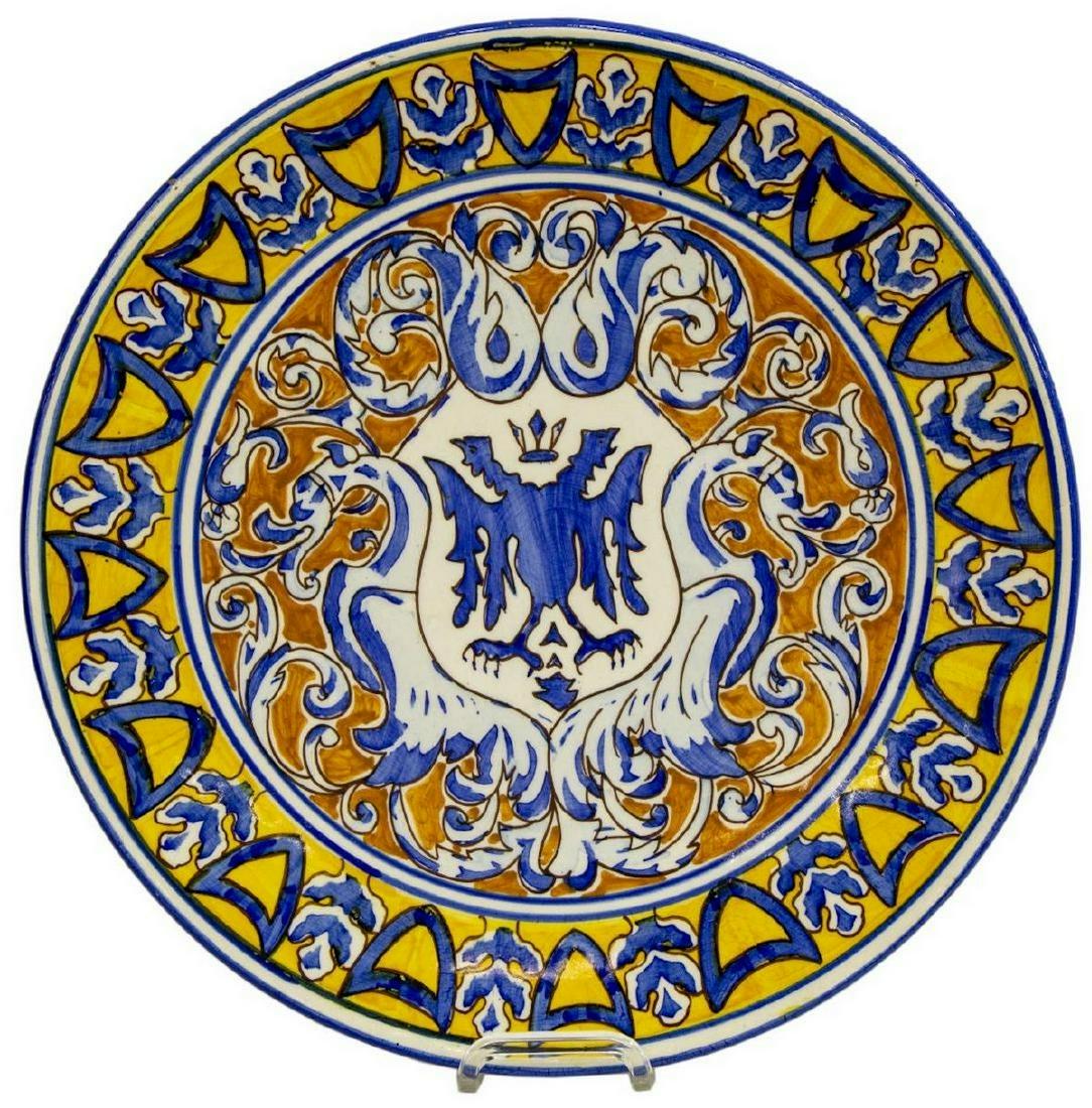 SPANISH MAJOLICA EMBLAZONED WALL CHARGER: Spanish majolica earthenware charger, early 20th c., a white ground decorated in blue and yellow, stylized border, enclosing a heraldic shield flanked by winged griffins, numbered to the back, short f