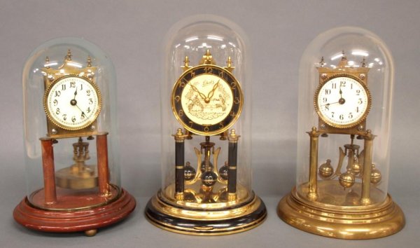 (3) GERMAN ANNIVERSARY CLOCKS: (lot of 3) German Anniversary clocks, all under glass domes: (1) August Schatz & Sohne, Germany, gilt and black finish with 18th century couple on face, metal base, 12"h; (1) porcelain face, mounted o