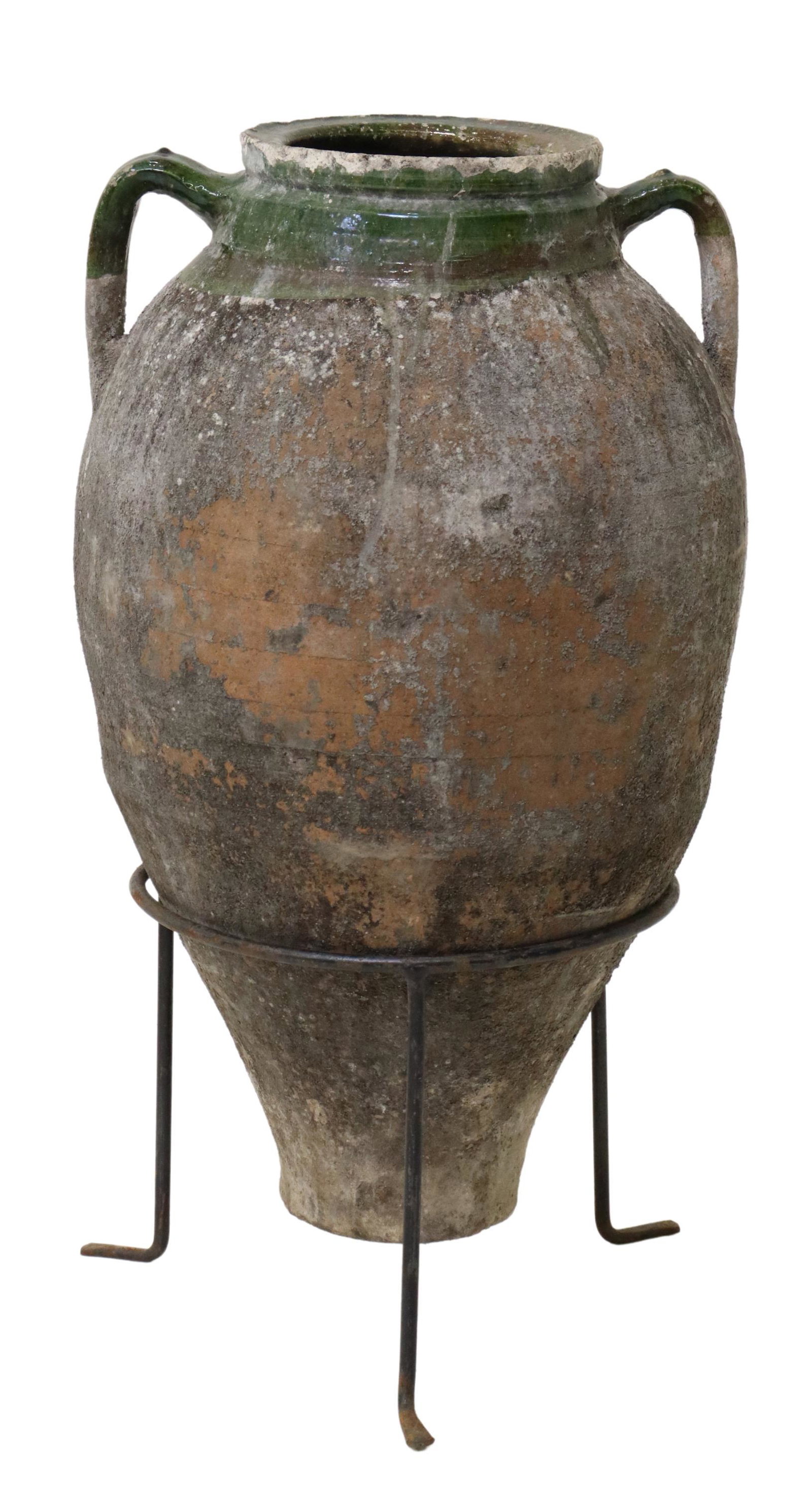 LARGE GREEN GLAZED EARTHENWARE OLIVE JAR IN STAND: Large earthenware olive jar in a green glaze, early 20th c., wide rim, over tapered body, with dual top handles, resting in iron tripartite base, approx 28.25"h, 15.75"diam Start Price: $200.00
