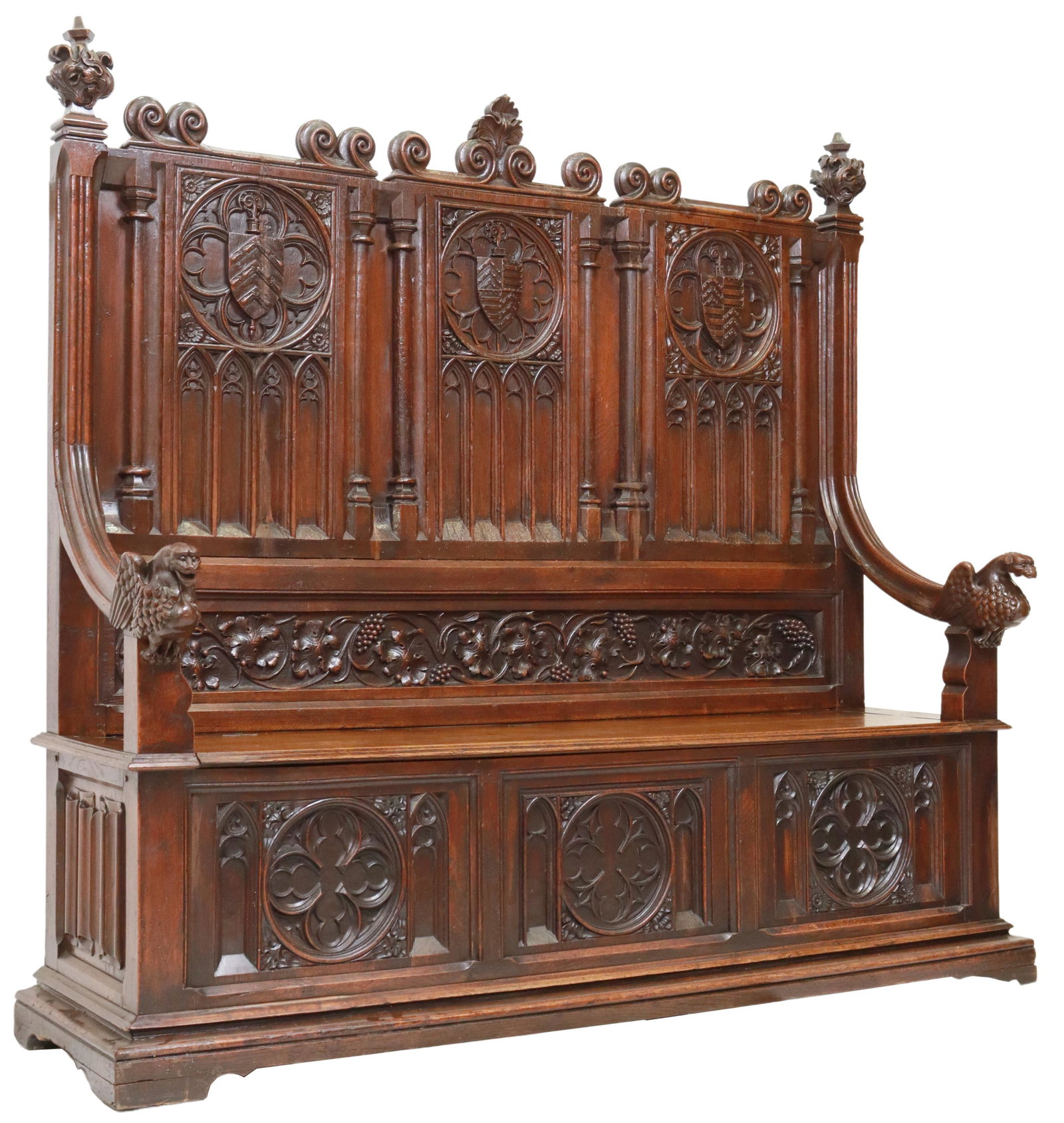 FRENCH GOTHIC REVIVAL CARVED ARMORIAL HALL BENCH: Fine French Gothic Revival oak hall bench, 19th c., scroll crest, over paneled backboard with coats of arms, pointed arches, hinged bench seat, arms terminating in carved winged grotesques, front pane