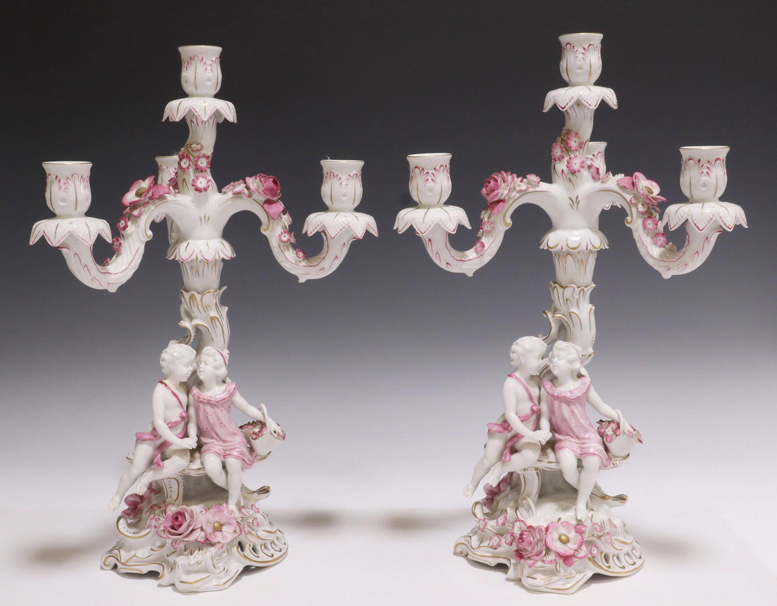 (2) DRESDEN STYLE FIGURAL PORCELAIN CANDELABRA: (pair) Dresden style parcel gilt porcelain candelabra, having four candle nozzles, over naturalistic tree-form standard, seated putto figure beside girl with basket of flowers, blue underglaze crowned