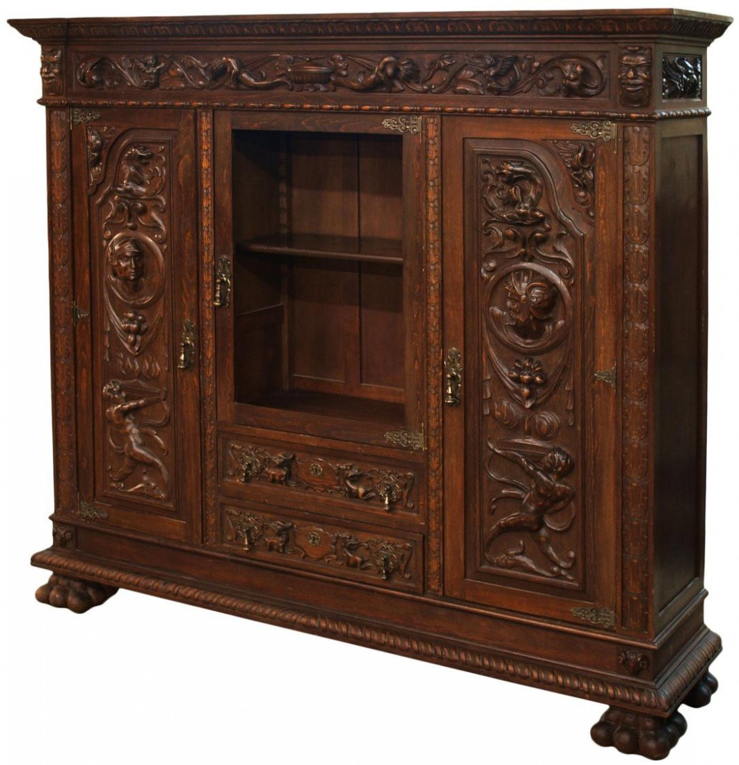 ANTIQUE SPANISH LIBRARY BOOKCASE: Antique Spanish bookcase, c. 1900, a stepped cornice over a figural and foliate carved frieze, a central glass door above two drawers, flanked by figural carved doors, plinth base raised by large doub