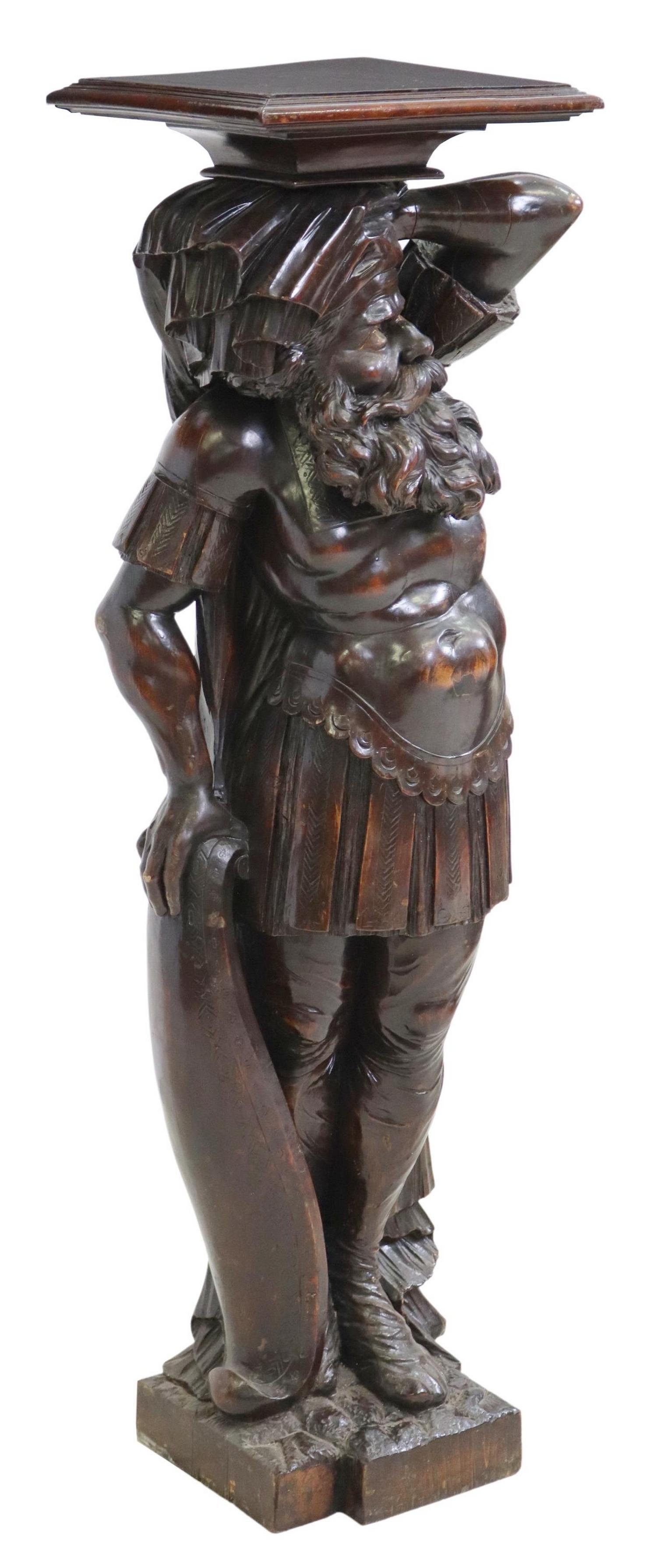 FIGURAL CARVED PEDESTAL STAND: Figural carved pedestal stand, 20th c., square top, over full-figured bearded man, dressed in stylized armor and holding a shield, rising on carved base, approx 51.75"h, 17"w, 14"d Start Price: $400.0