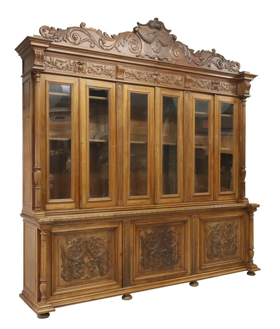 MONUMENTAL FRENCH CARVED WALNUT BOOKCASE: Monumental French carved walnut bookcase, early 20th c., having large scrolling foliate crest, centering shield, six glazed doors, flanked by fluted and turned side supports, over lower case fitted wi