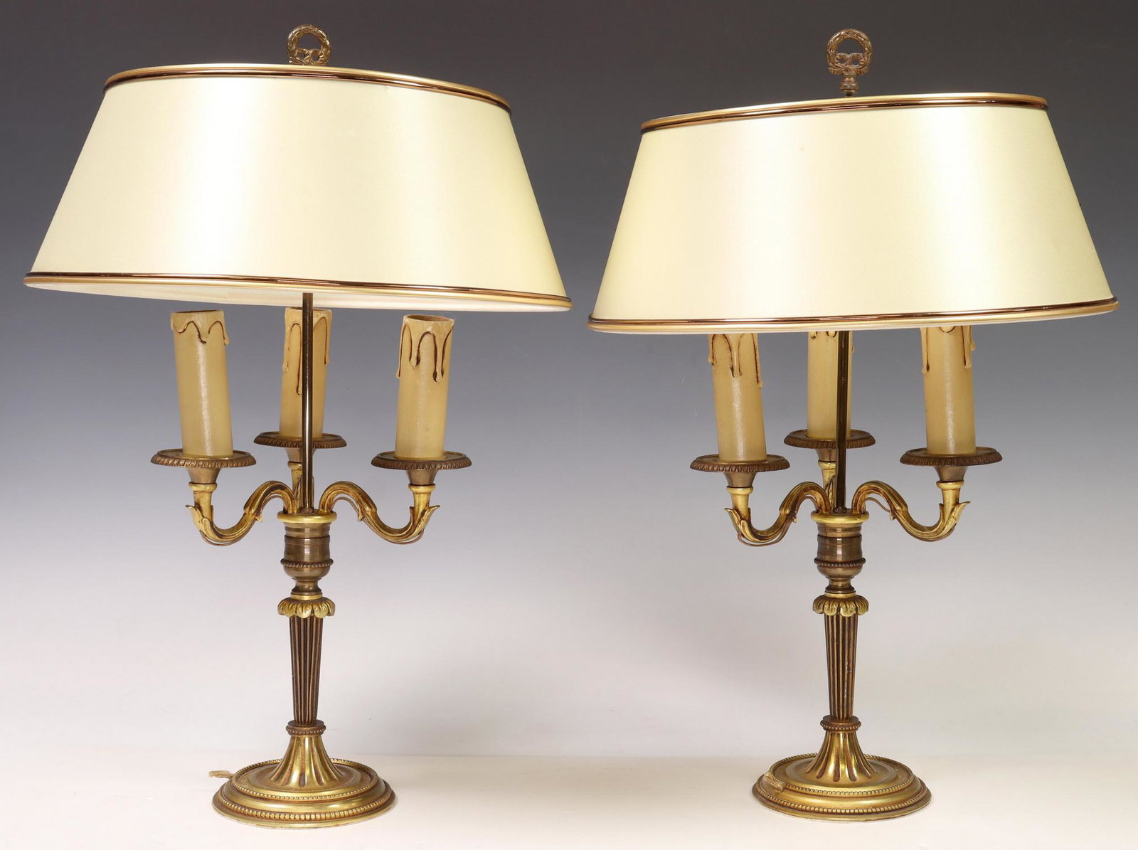 (2) FRENCH LOUIS XVI STYLE BOUILLOTTE 3-LT LAMPS: (pair) French Louis XVI style gilt metal bouillotte lamps, 20th c., cream shades, over fluted and tapered standard, issuing foliated arms, terminating in three lights, rising on round beaded base, in 